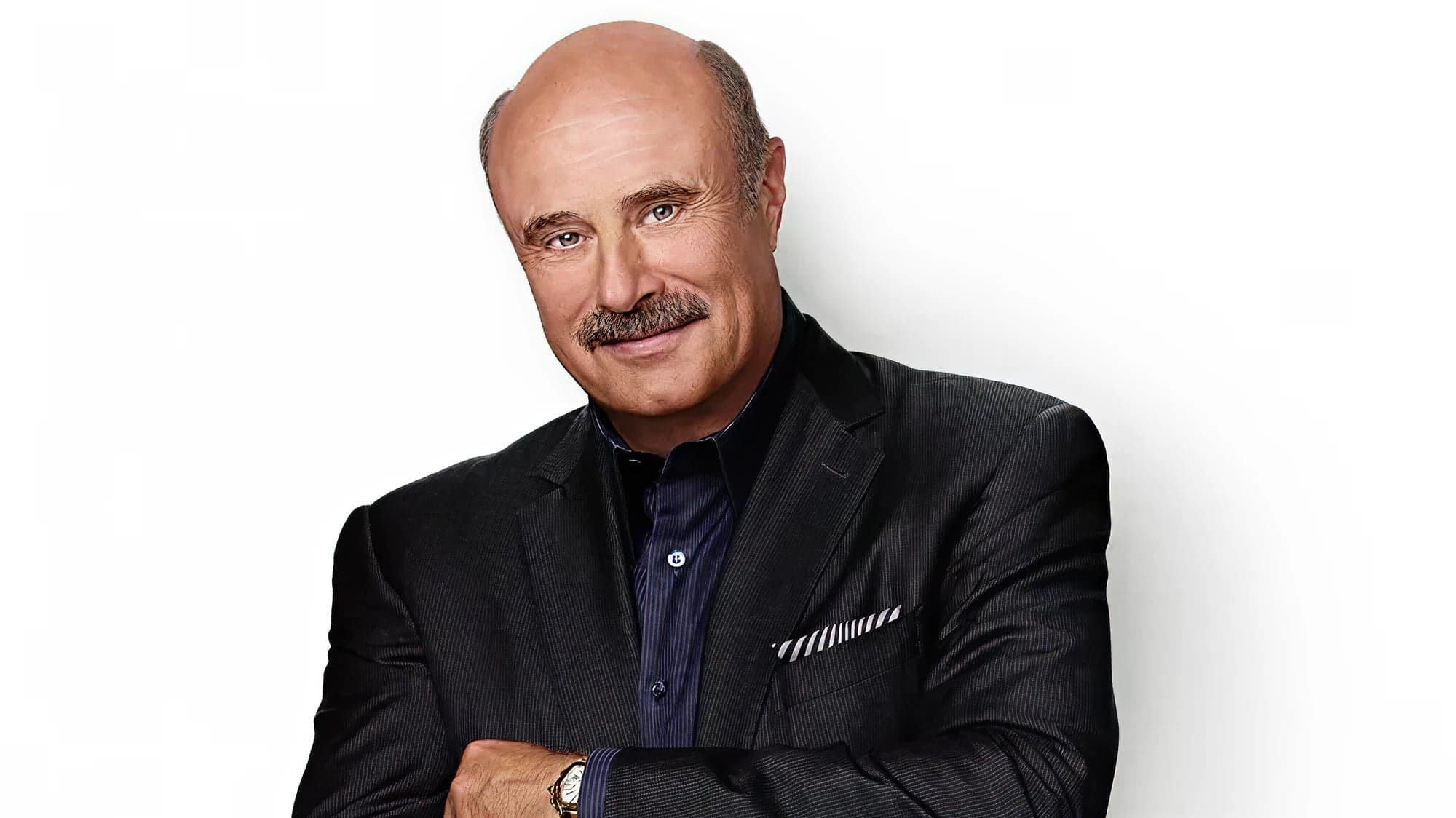 Dr. Phil McGraw — arms folded, in a dark suit, against a white studio background