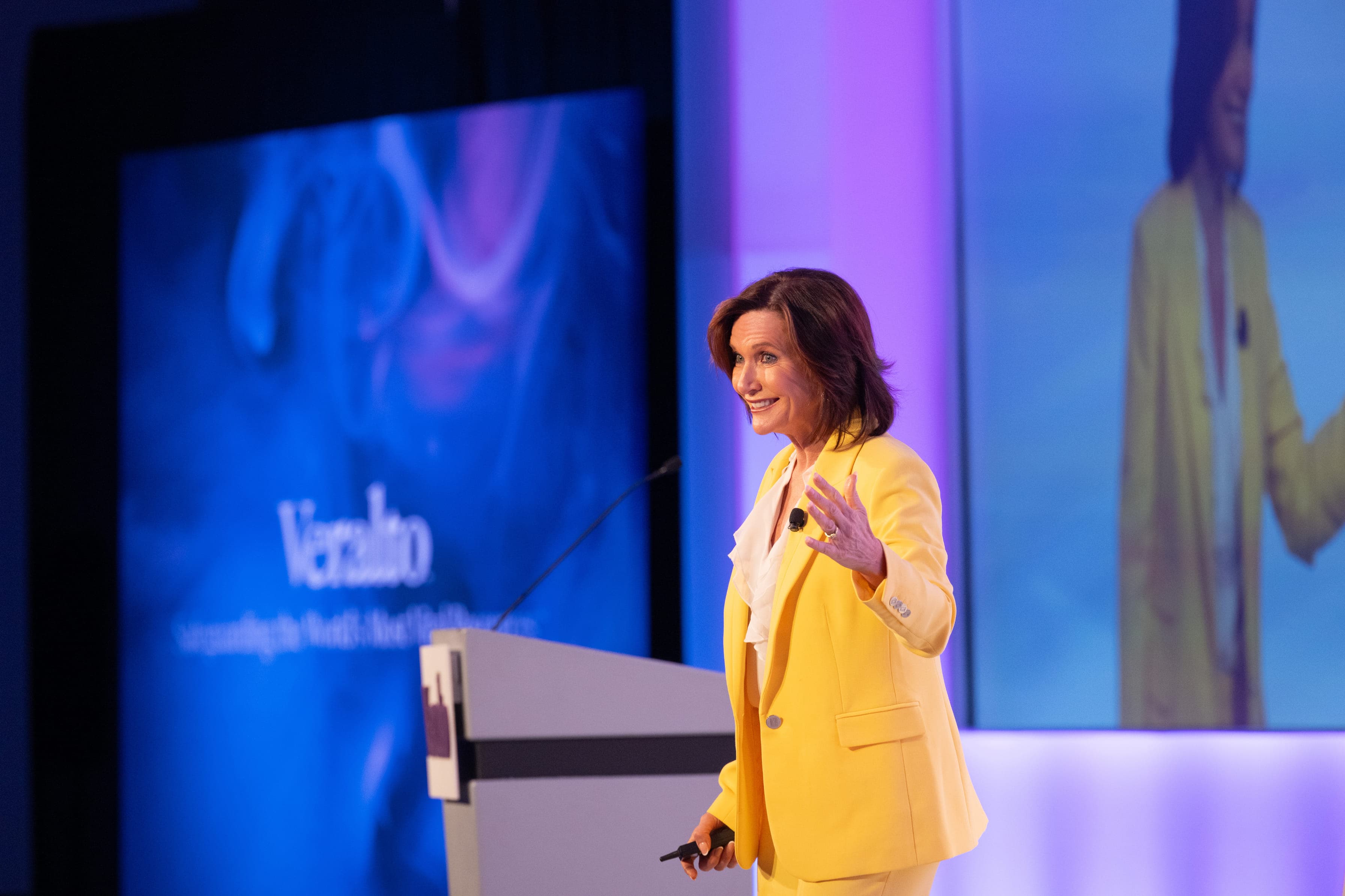 Dr. Margie Warrell speaking on a conference stage in a bright yellow blazer over a cream blouse, her right hand raised open-palmed mid-gesture, in front of a large blue-to-purple LED screen