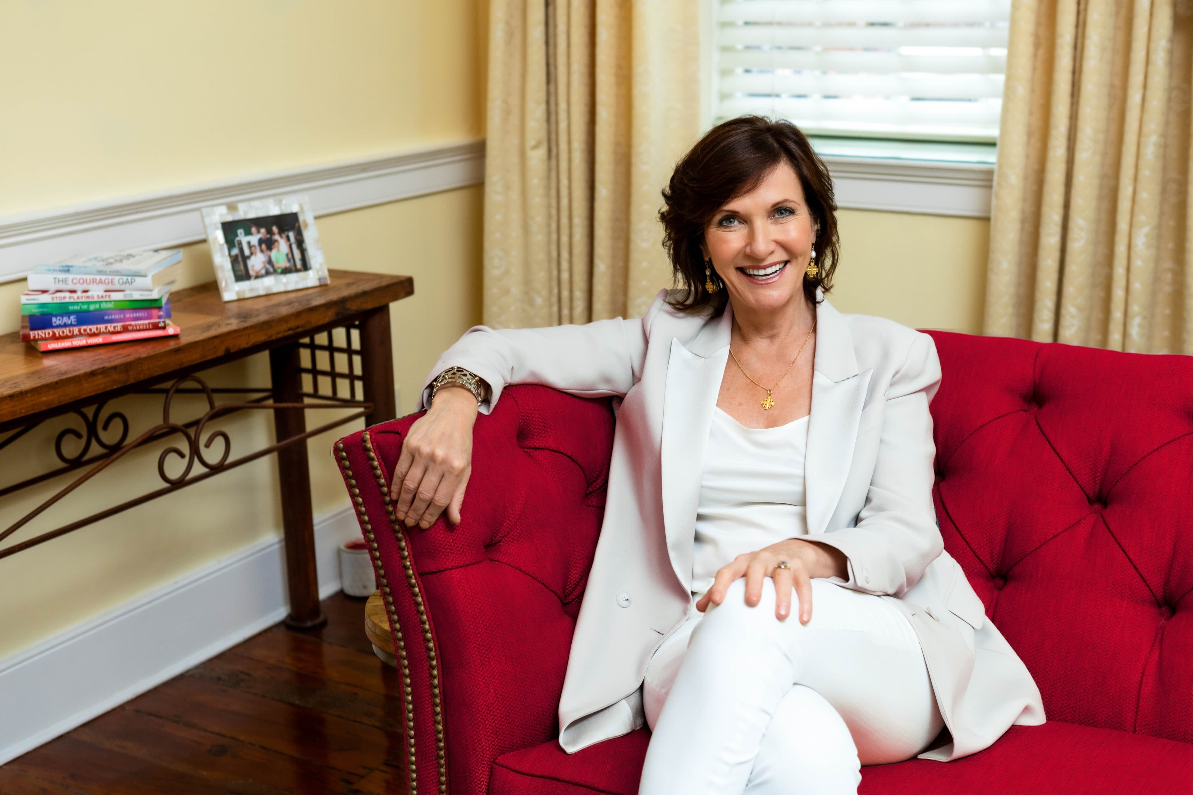 Dr. Margie Warrell seated on a crimson tufted armchair in a warm panelled living room, wearing a cream blazer and gold jewellery, smiling to camera