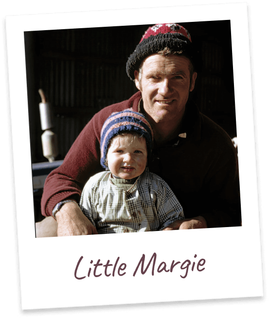 Little Margie. Margie's father in a knitted beanie and dark red jumper holding her as a toddler inside a farm shed.
