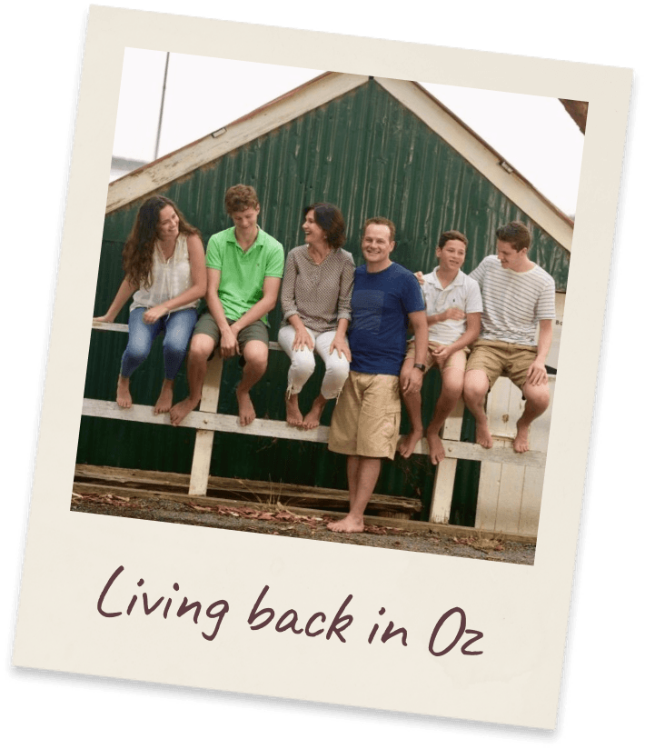 Living back in Oz. Six young people sitting barefoot in a row along a white timber fence rail in front of a green corrugated-iron barn.