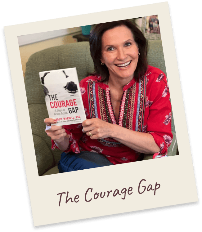 The Courage Gap. Margie seated in a green armchair in a red-and-white patterned blouse, holding up her book The Courage Gap.
