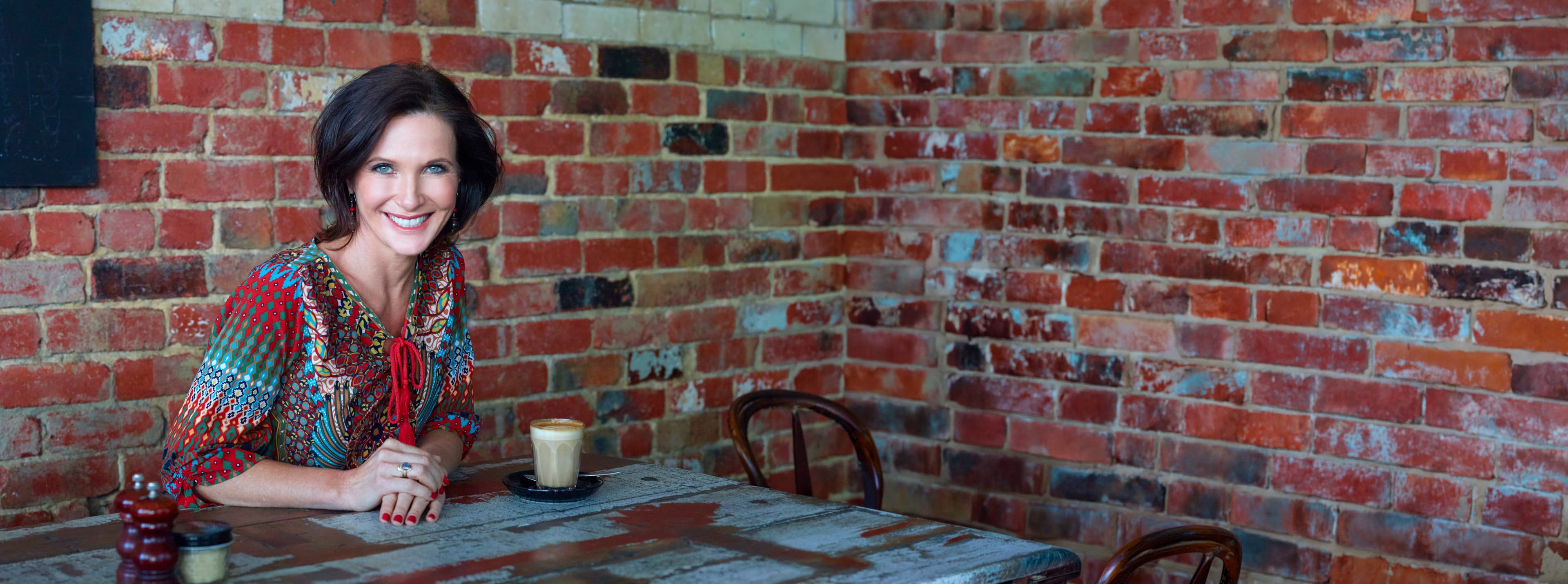 Dr. Margie Warrell seated at a café table in front of a dark red-brown brick wall, smiling to camera in a patterned teal, red and gold blouse with a red tassel tie, a latte in a glass on the table in front of her