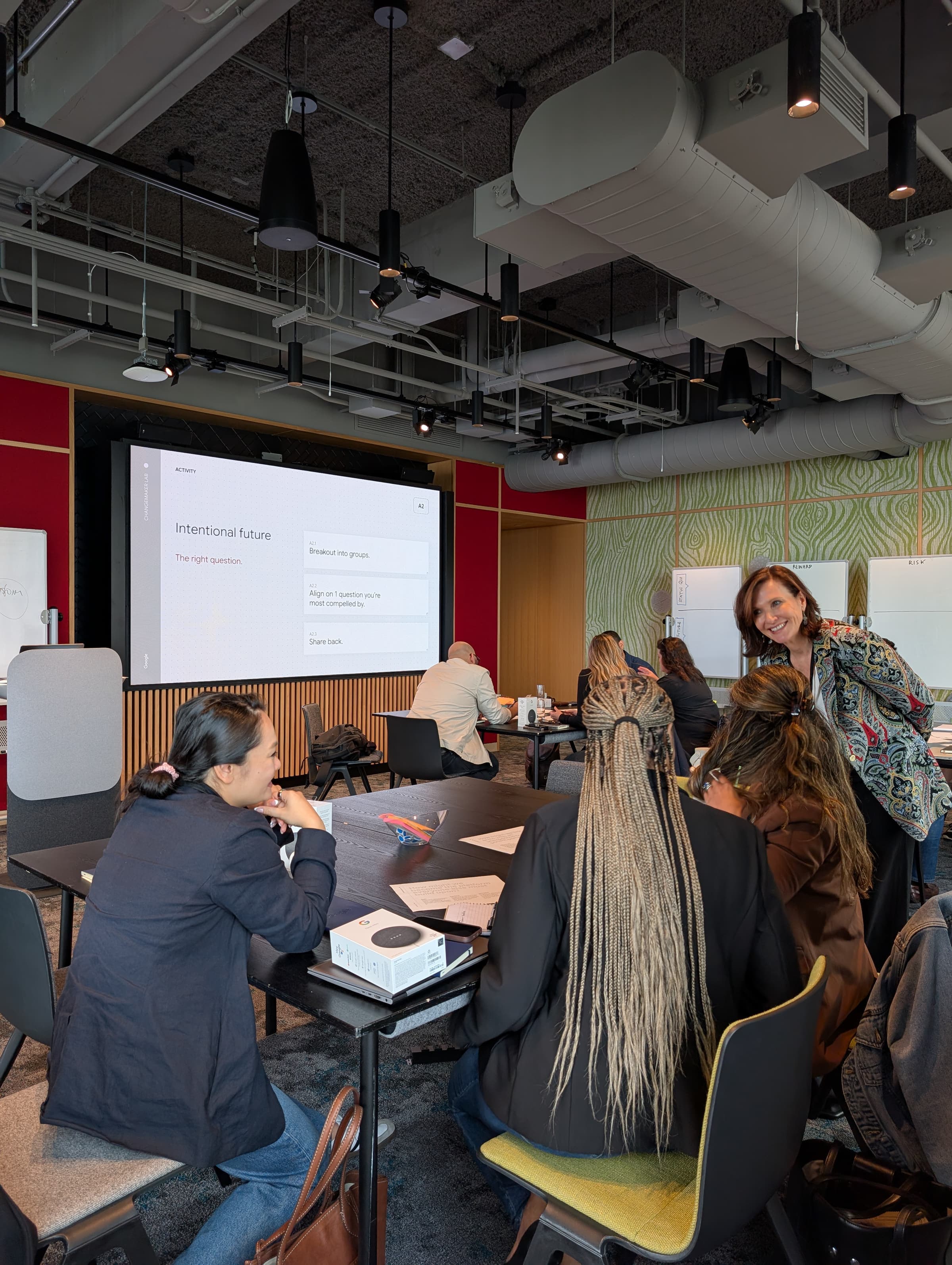 Dr. Margie Warrell laughing with a table of participants at a Courage Lab session, leaning in beside their desk while the room’s wall screen shows the Lab’s “Intentional future — the right question” activity slide and sticky-note whiteboards line the wall behind her