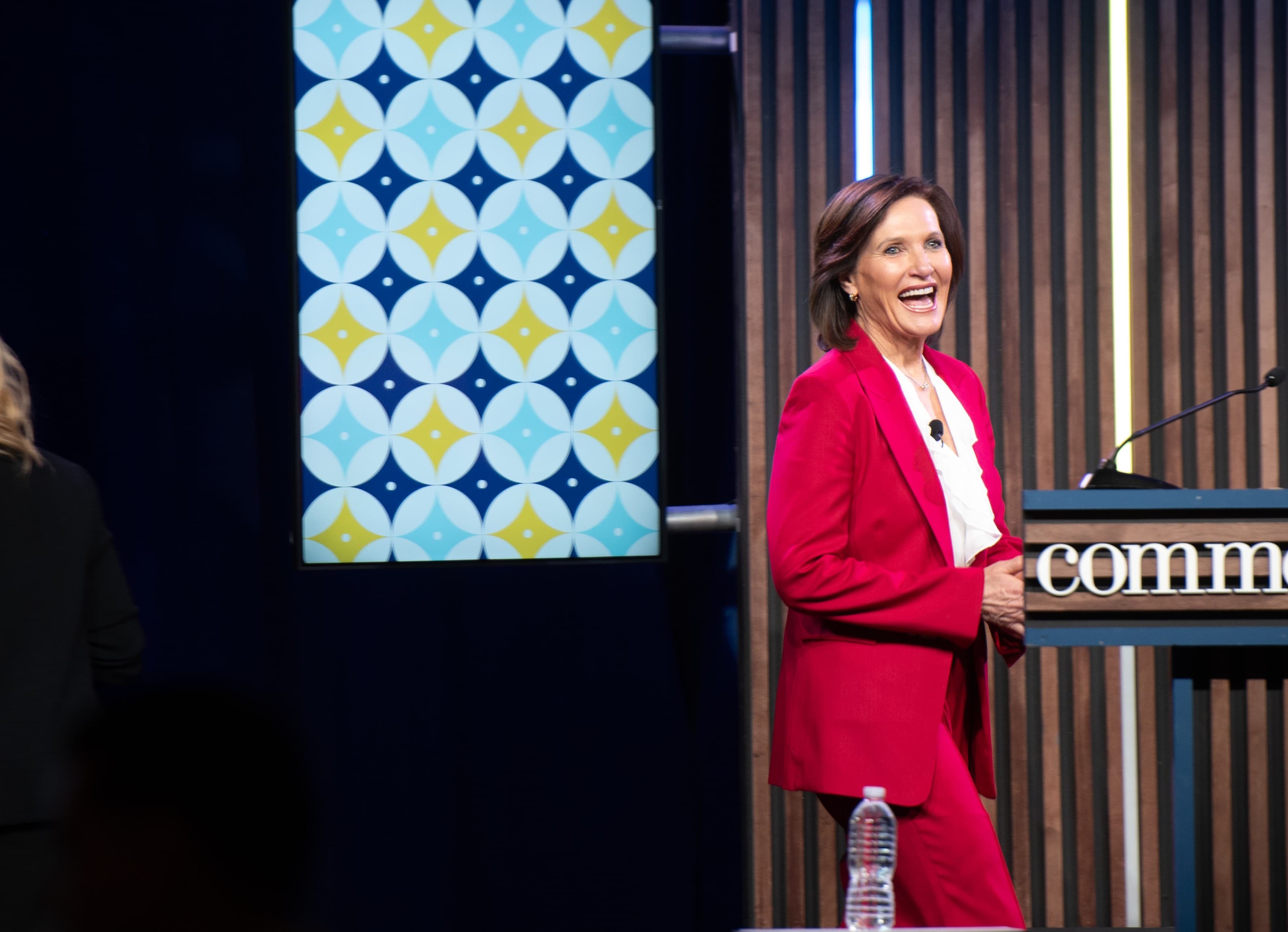 Dr. Margie Warrell laughing on stage in a magenta-red suit beside a slatted timber lectern, a tall patterned banner to her left