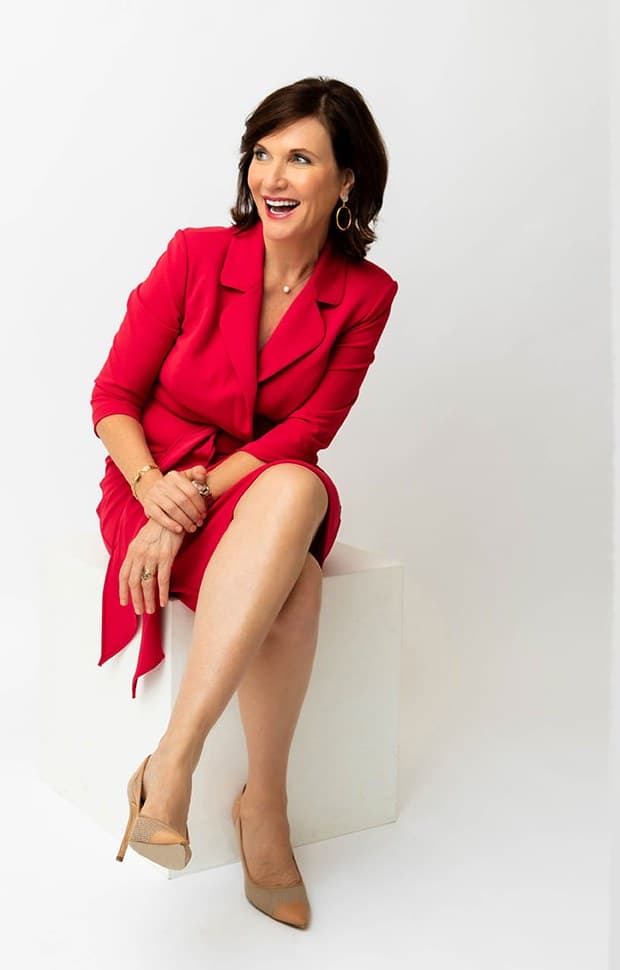 Dr. Margie Warrell seated on a white block in a red wrap dress and nude heels, laughing and looking off to one side