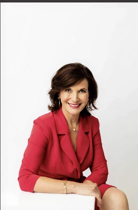 Dr. Margie Warrell in a crimson blazer and matching dress, seated at a white table with her hands lightly crossed, against a blown-out white studio wall