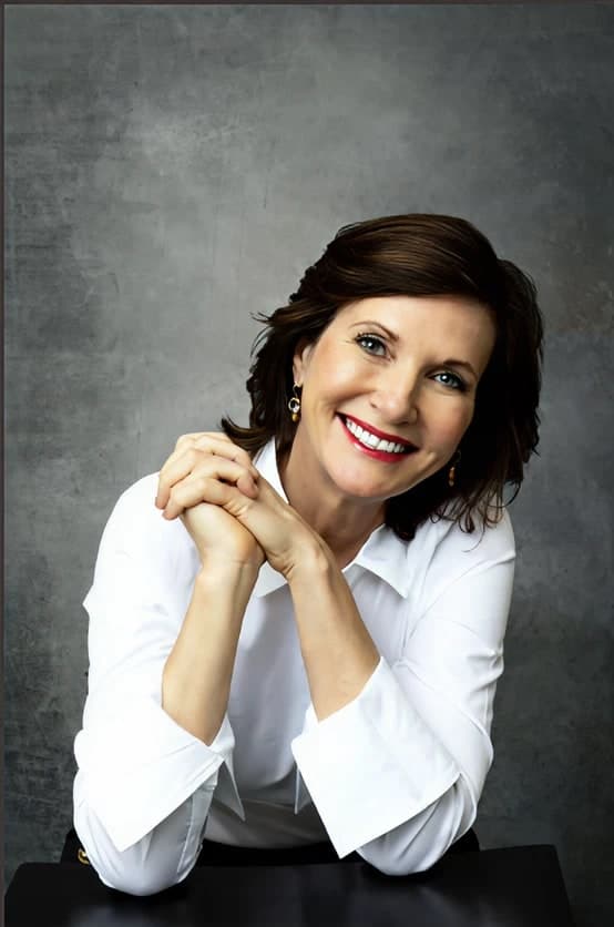 Dr. Margie Warrell in a crisp white shirt with turned-back cuffs, leaning on her elbows with fingers interlaced under her chin, smiling widely against a mottled charcoal backdrop