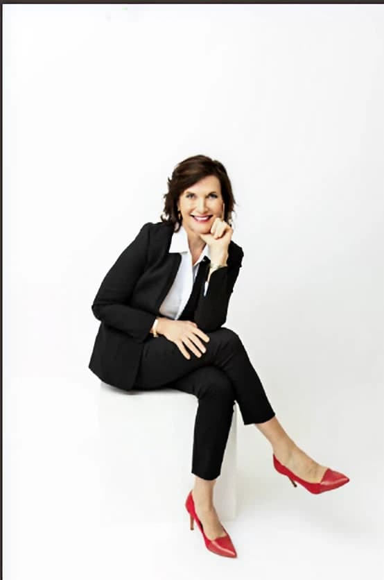 Dr. Margie Warrell full length in a black trouser suit over a white shirt with red pointed heels, perched on a low white block with her legs crossed, on a high-key white background