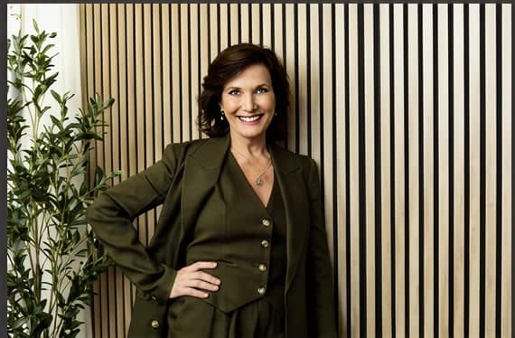 Dr. Margie Warrell in an olive-green gold-buttoned jacket and skirt, one hand on her hip, standing in front of a vertical timber batten wall