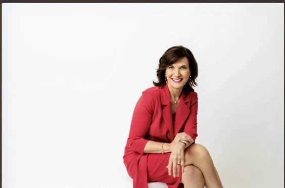 Dr. Margie Warrell in a red blazer and dress, seated right of centre with her legs crossed, in a wide and largely empty pale studio frame
