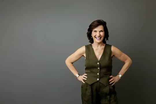 Dr. Margie Warrell laughing in a sleeveless olive waistcoat and matching trousers, both hands on her hips, against a flat olive-grey studio backdrop