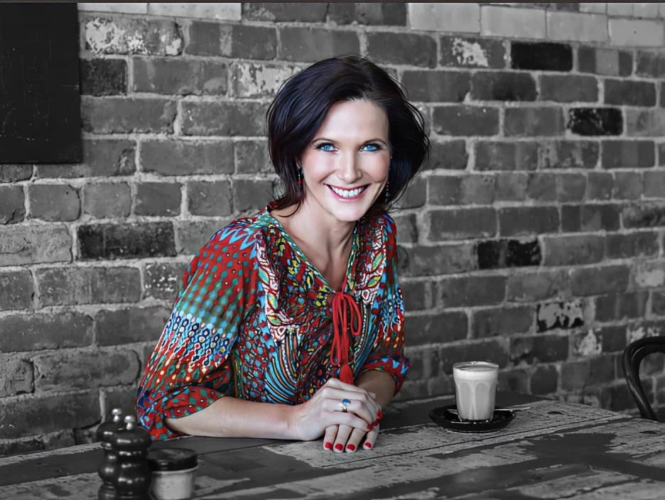 Dr. Margie Warrell leaning on her forearms over a weathered timber café table in a multicoloured geometric-print blouse with a red tie neck, shot in colour against a desaturated brick wall