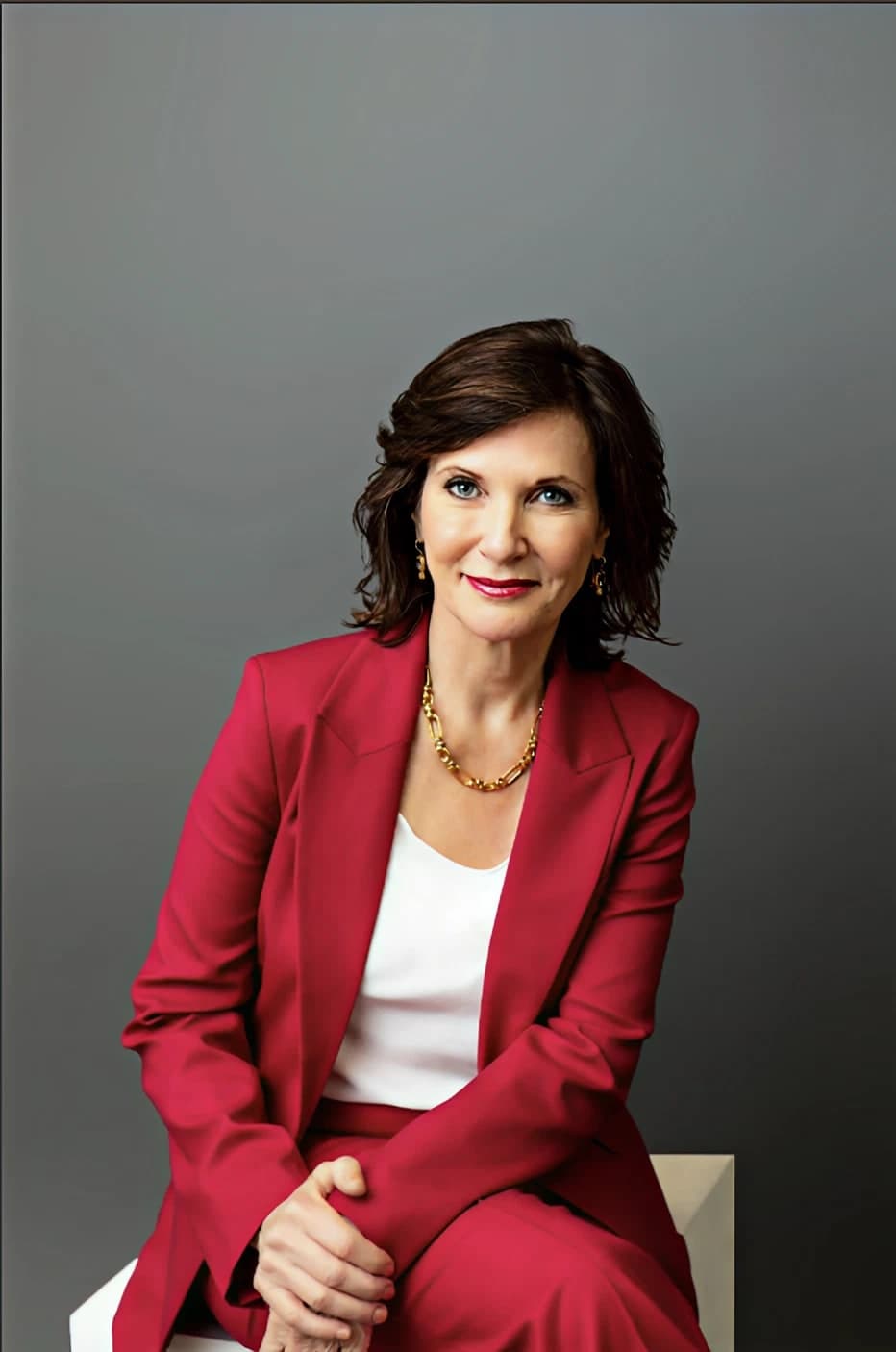 Dr. Margie Warrell waist-up on a cream stool in a crimson blazer over a white top with a chunky gold collar necklace, hands clasped in her lap, on a graduated mid-grey backdrop