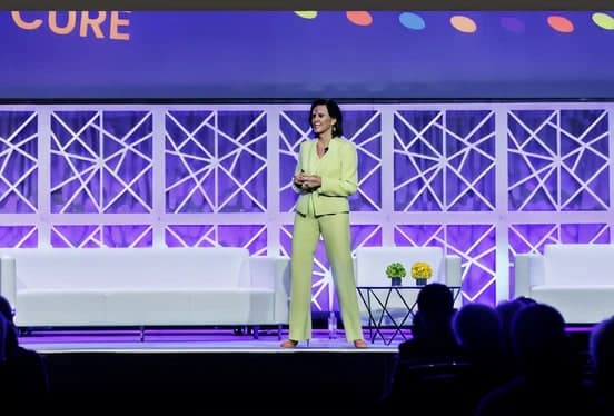 Dr. Margie Warrell on a white stage in a pale lime trouser suit, hands together, in front of a lattice-truss set piece washed in violet light with silhouetted audience heads in the foreground