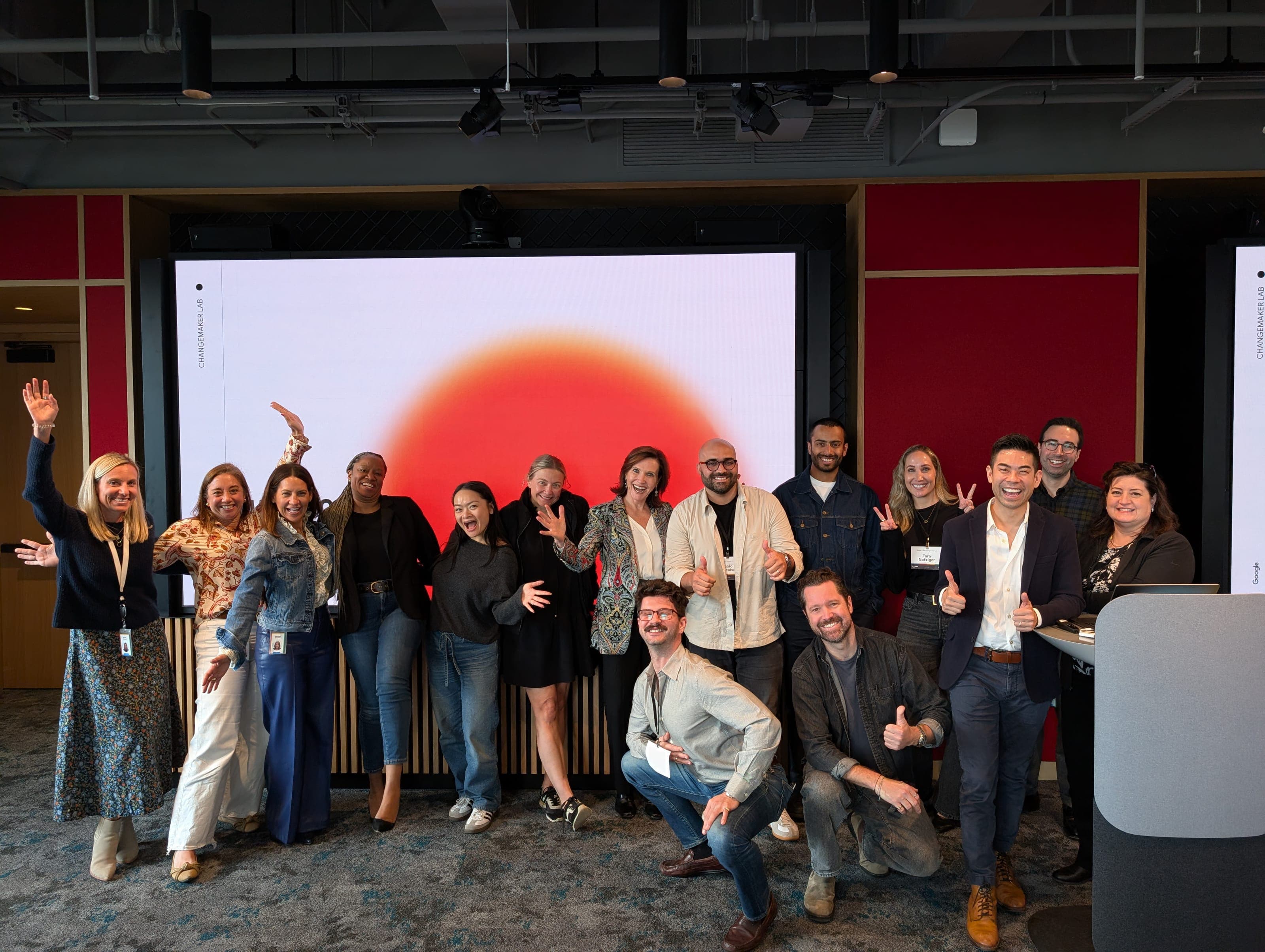 Around twenty people posed and grinning in a corporate event room, waving and giving thumbs-up, with Dr. Margie Warrell centre-left in a teal paisley jacket; behind them a wall screen filled with a large orange-red rising-sun graphic beside deep-red acoustic panels