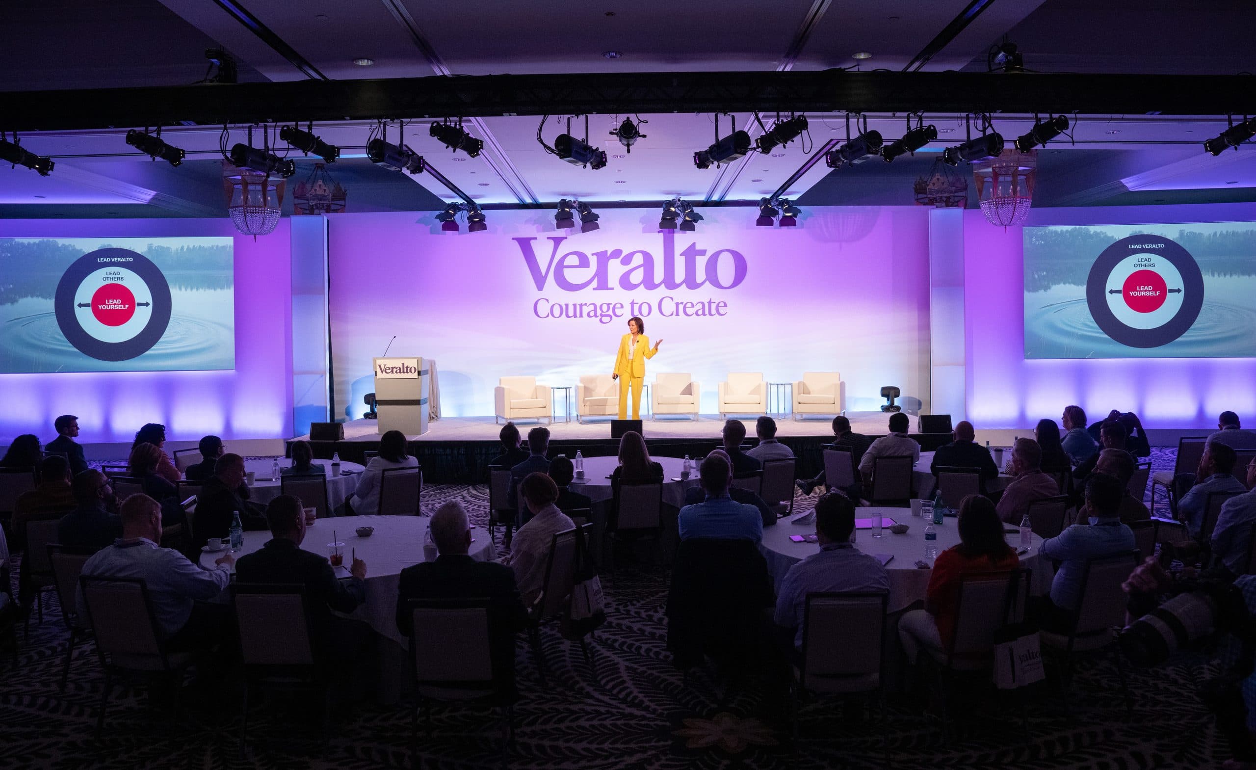 Dr. Margie Warrell in a yellow suit speaking on a ballroom stage at a Veralto leadership summit, "Courage to Create" projected behind her and a seated audience at round tables in the foreground