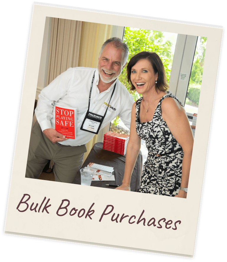 Polaroid captioned “Bulk Book Purchases”: Dr. Margie Warrell laughing in a black-and-white floral dress beside a grey-bearded man in a conference lanyard holding up a copy of Stop Playing Safe, with a stack of the same books on the table in front of them