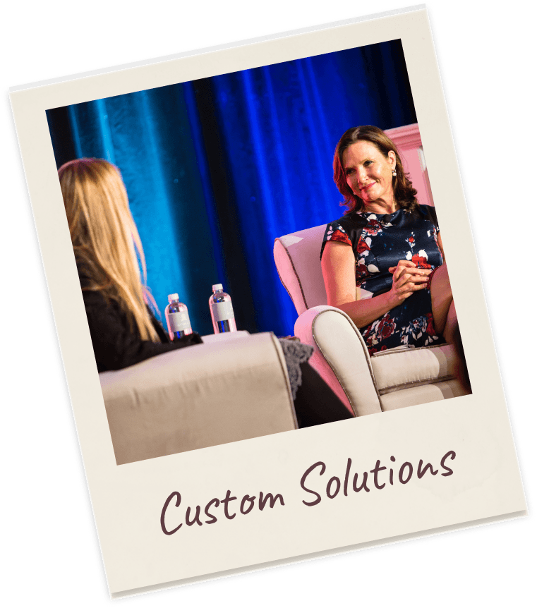Polaroid captioned “Custom Solutions”: Dr. Margie Warrell in a navy floral dress seated cross-legged in a cream armchair on stage under blue light, mid-conversation with an interviewer seated opposite