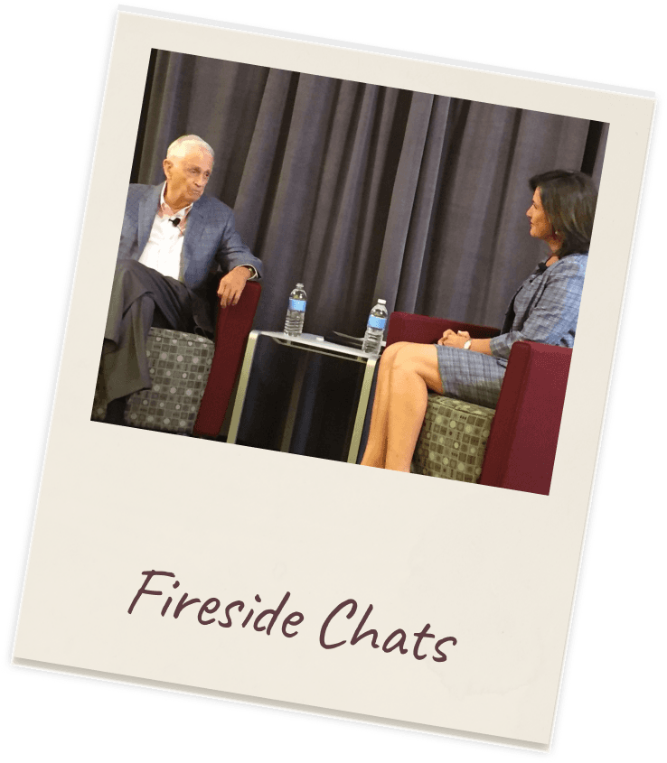 Polaroid captioned “Fireside Chats”: a two-armchair fireside-chat set with an older grey-haired man in a grey blazer at left and Dr. Margie Warrell listening at right in a pale-blue blazer, a small side table between them against a heavy dark curtain