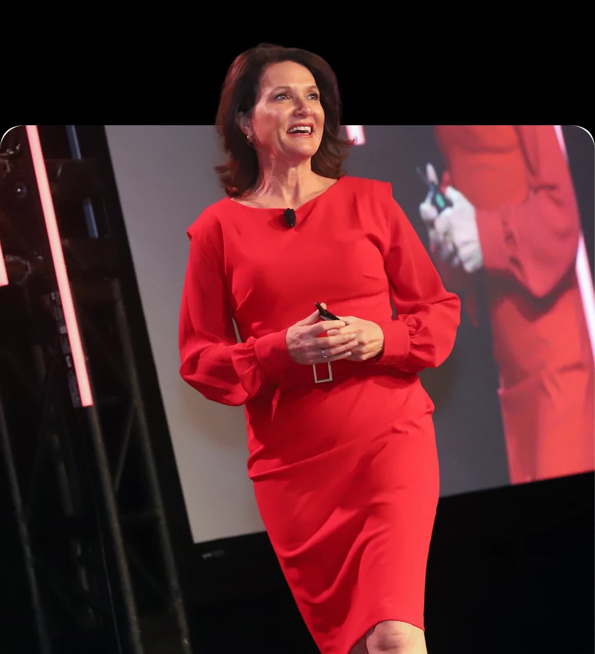 Dr. Margie Warrell speaking on stage in a red dress, a projection screen and stage truss behind her
